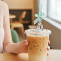 Hand holding a cup of iced coffee with a decorative straw and tag in a casual setting.