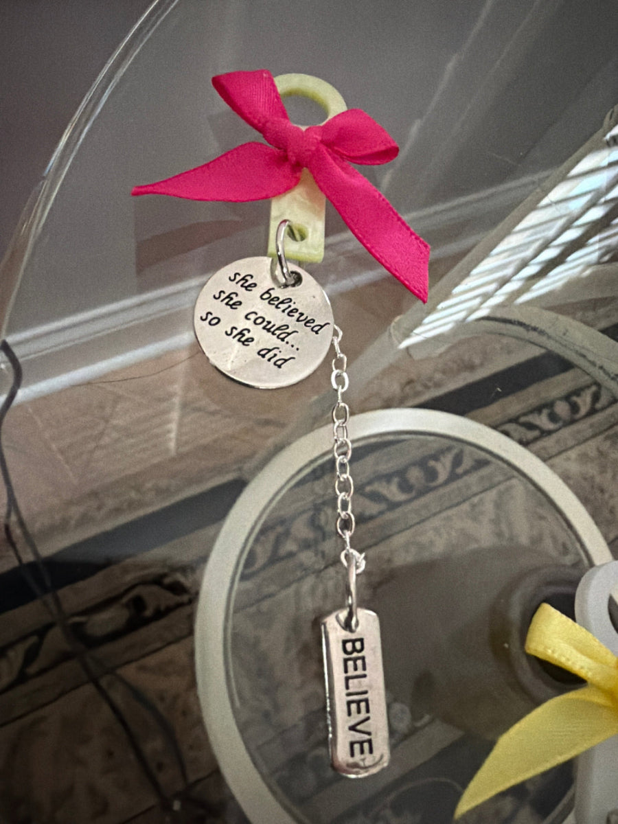 Decorative keychain with pink bow and 'BELIEVE' tag on a reflective surface